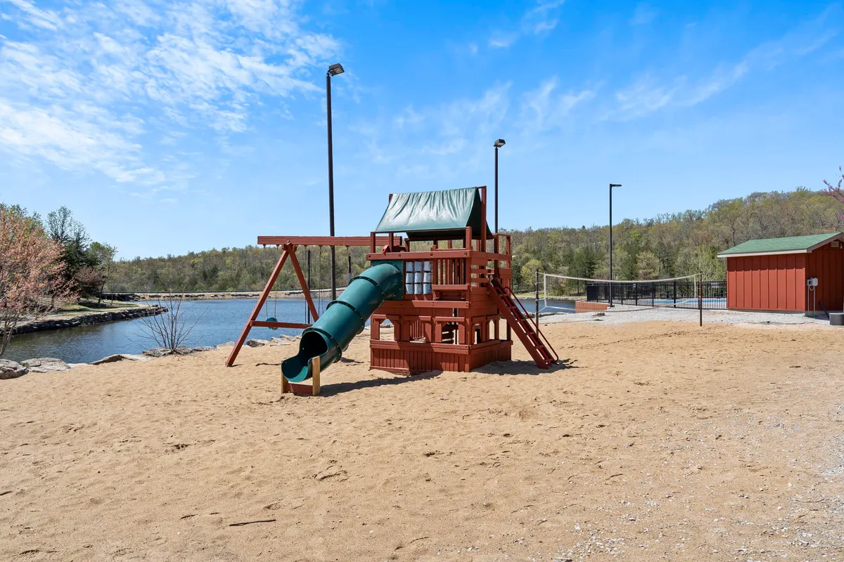 A vibrant playground area featuring a colorful slide, sand pit, water body in the background, and surrounded by lush green trees. Children can be seen playing and families are gathered around for picnics and outdoor fun.