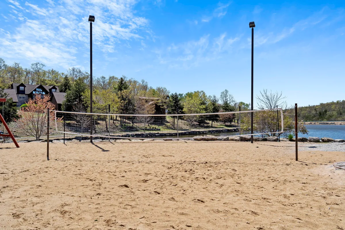 A sand volleyball court is set near a tranquil lake, surrounded by trees with playground equipment visible nearby. Families and groups are enjoying volleyball games, picnicking by the water, and outdoor recreation in a scenic resort setting.