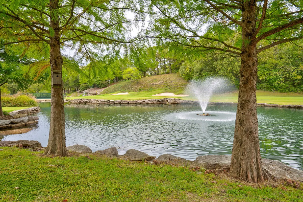 A tranquil golf course scene centered around a serene fountain, bordered by water, green fairways, and mature trees. The peaceful mood invites relaxation and moments of post-game reflection.