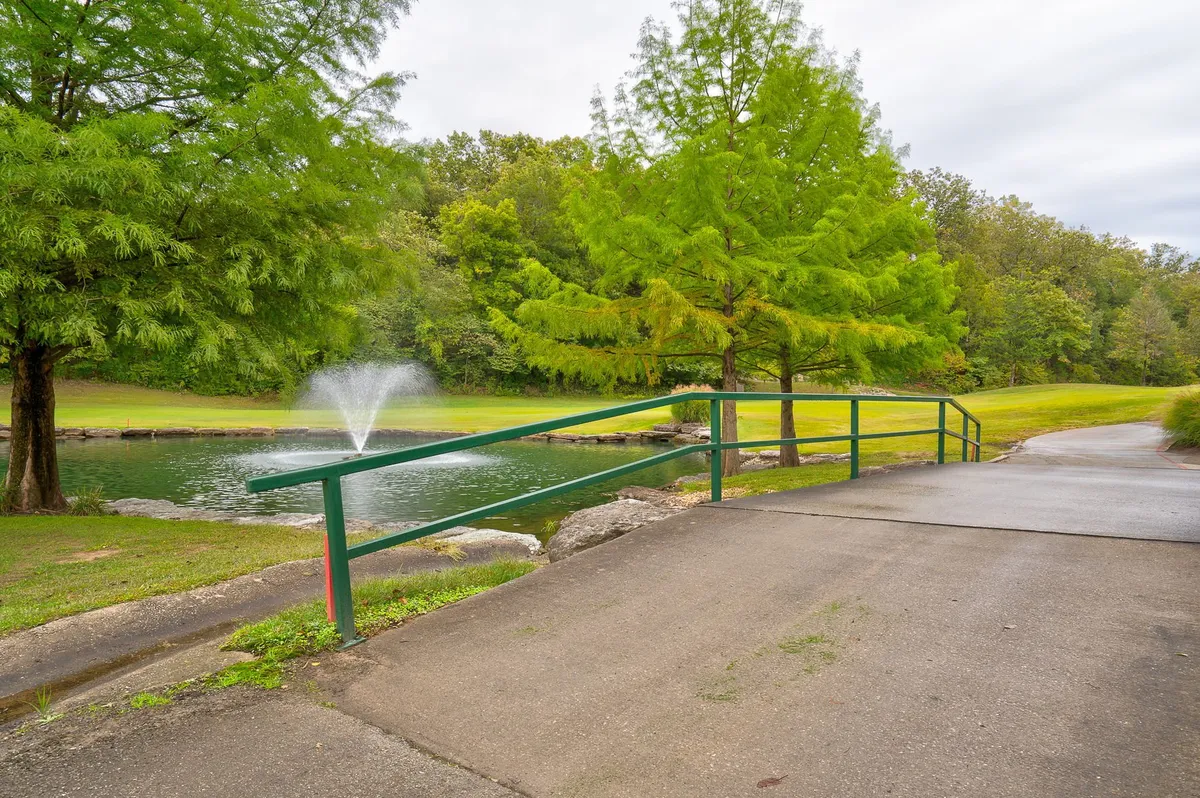 A serene pond with a dancing fountain framed by green railings and trees creates a peaceful recreational spot. The tranquil setting is perfect for fishing, walking, and enjoying nature within the resort grounds.