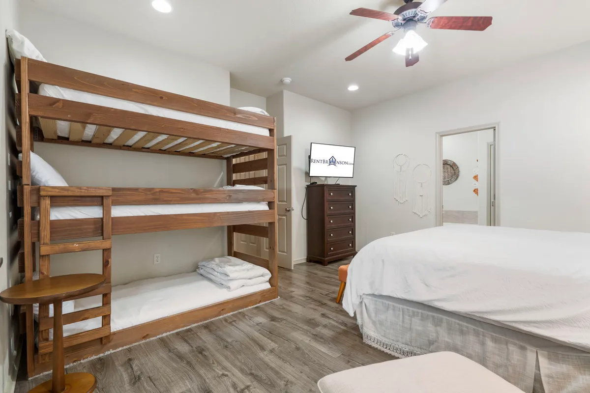 The image showcases a bedroom furnished with both a queen bed and a cozy bunk bed. There's also a TV and dresser, making the room practical for families or groups. The triple twin bunk bed setup is particularly inviting for young guests.