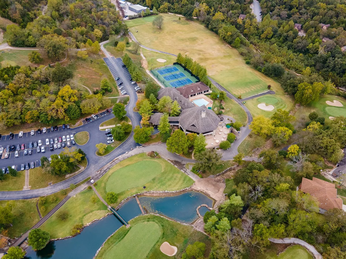 A broad exterior shot capturing the Ledgestone Golf Course with its manicured greens, tennis courts, main building, pond, and ample parking. The image is framed by lush landscaping and shows golfers and visitors enjoying the amenities.
