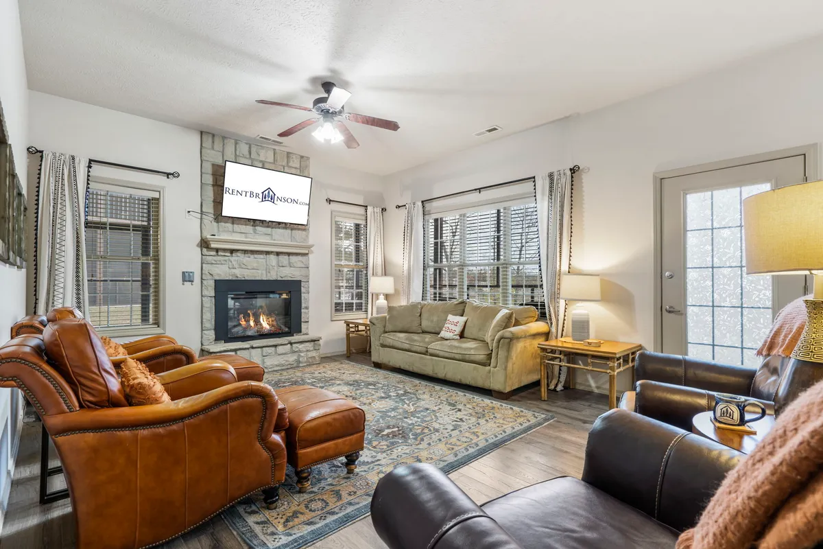 The image shows a cozy living room with a welcoming fireplace as the centerpiece, surrounded by a comfortable sofa and plush recliners. Soft light filters in, creating a warm atmosphere perfect for relaxing, watching TV, or gathering with family and friends. The rug and tasteful decor add to the inviting ambiance, making it an ideal spot for quiet mornings with coffee or memorable game nights.